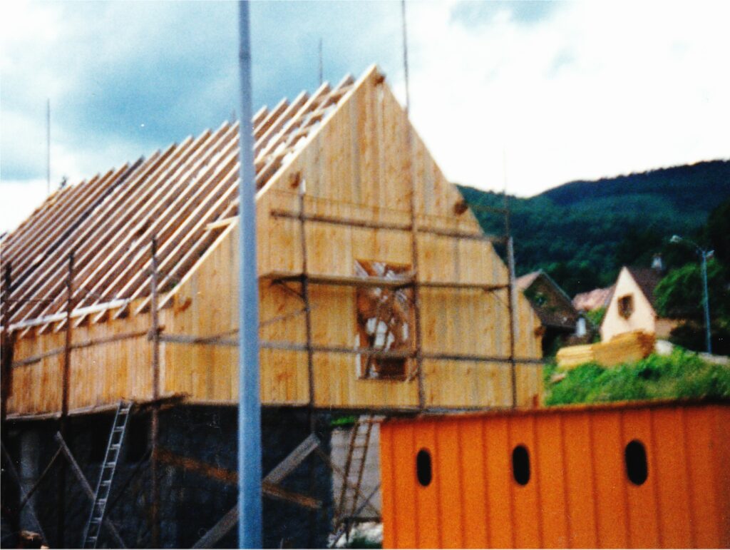 Under-construction wooden house with exposed roof rafters; orange construction barrier in the foreground and hills in the distance.