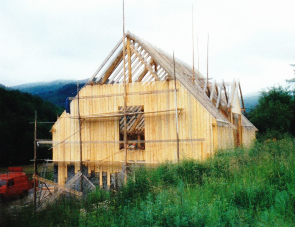 Wood-framed house under construction with exposed roof trusses and scaffolding in a grassy, rural field. Mountains in the background.