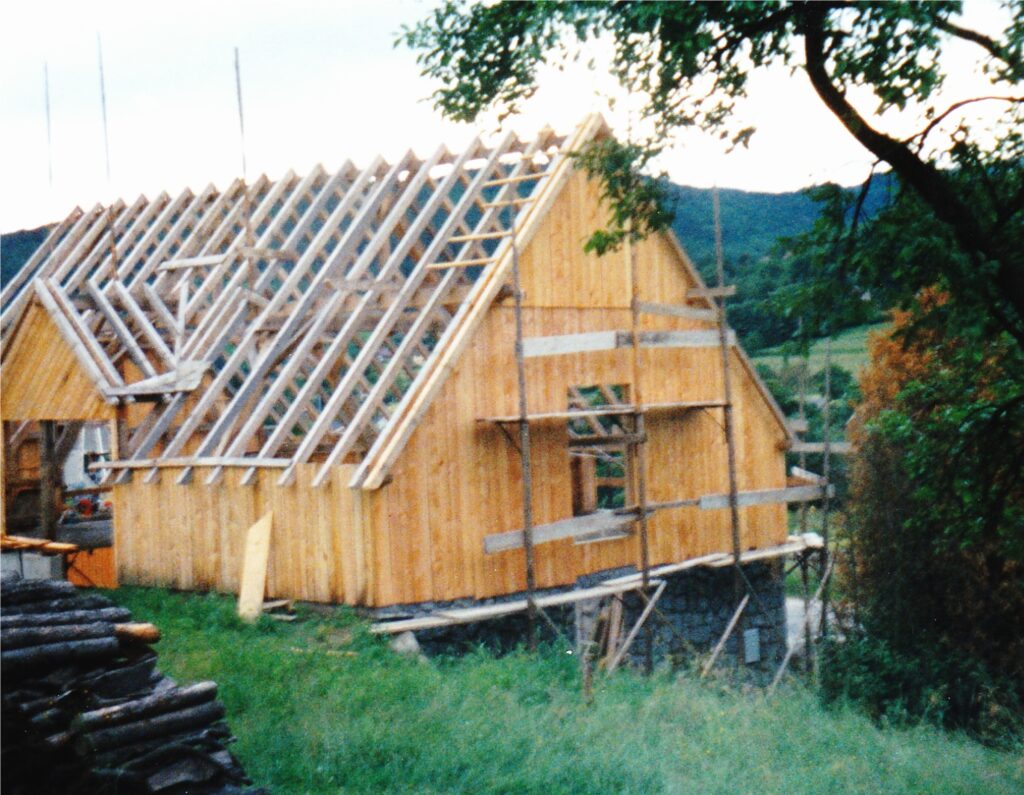 Wood-framed house under construction with exposed roof trusses and scaffolding, set in a green hillside landscape