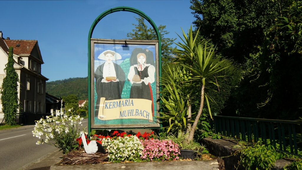 Roadside sign with a folk-art painting of a man and woman in traditional dress under a green arch, displaying the text 'KERMARIA MUHLBACH' amid a flower bed.