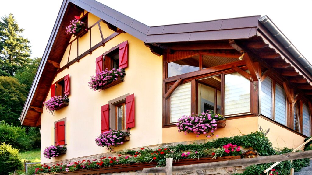 Yellow cottage-style house with red shutters and pink flower boxes along the windows.