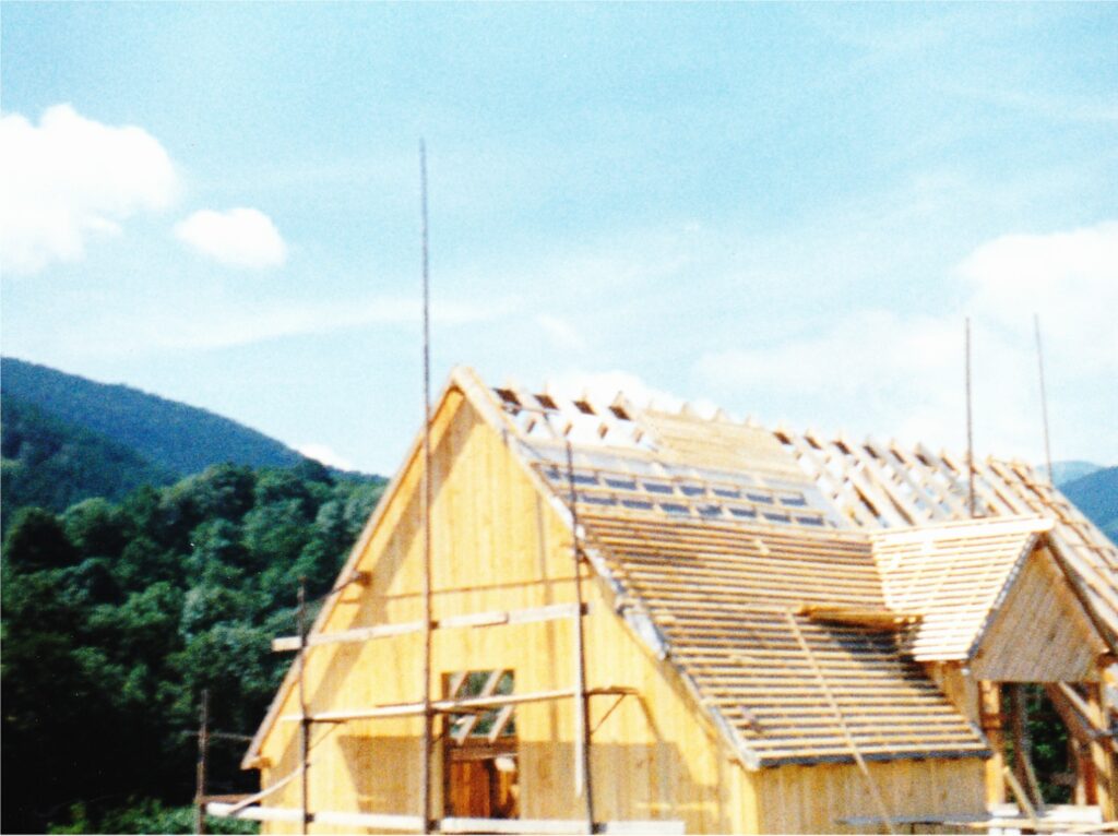 Wooden house frame under construction with exposed roof trusses and scaffolding, set in a mountainous countryside under a blue sky.