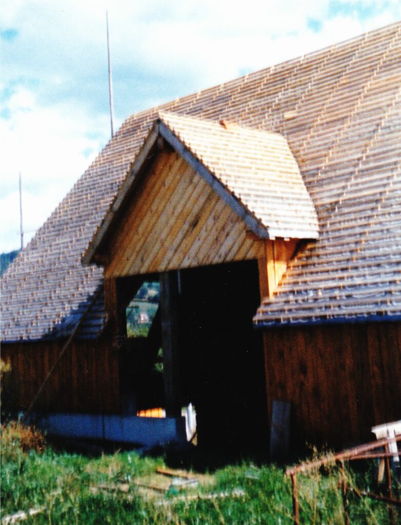 Wooden house under construction with a steep gabled roof and an open doorway opening in the front.