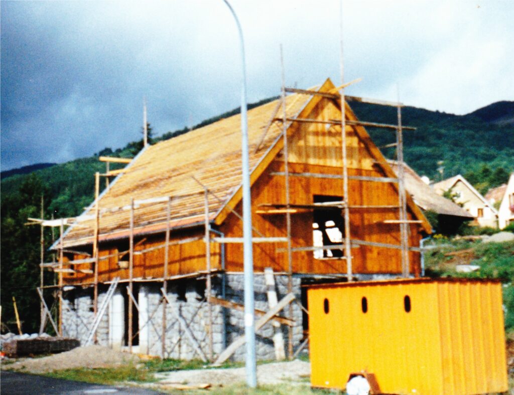 Wood-framed house under construction with scaffolding on a hillside and a yellow storage container in the foreground.