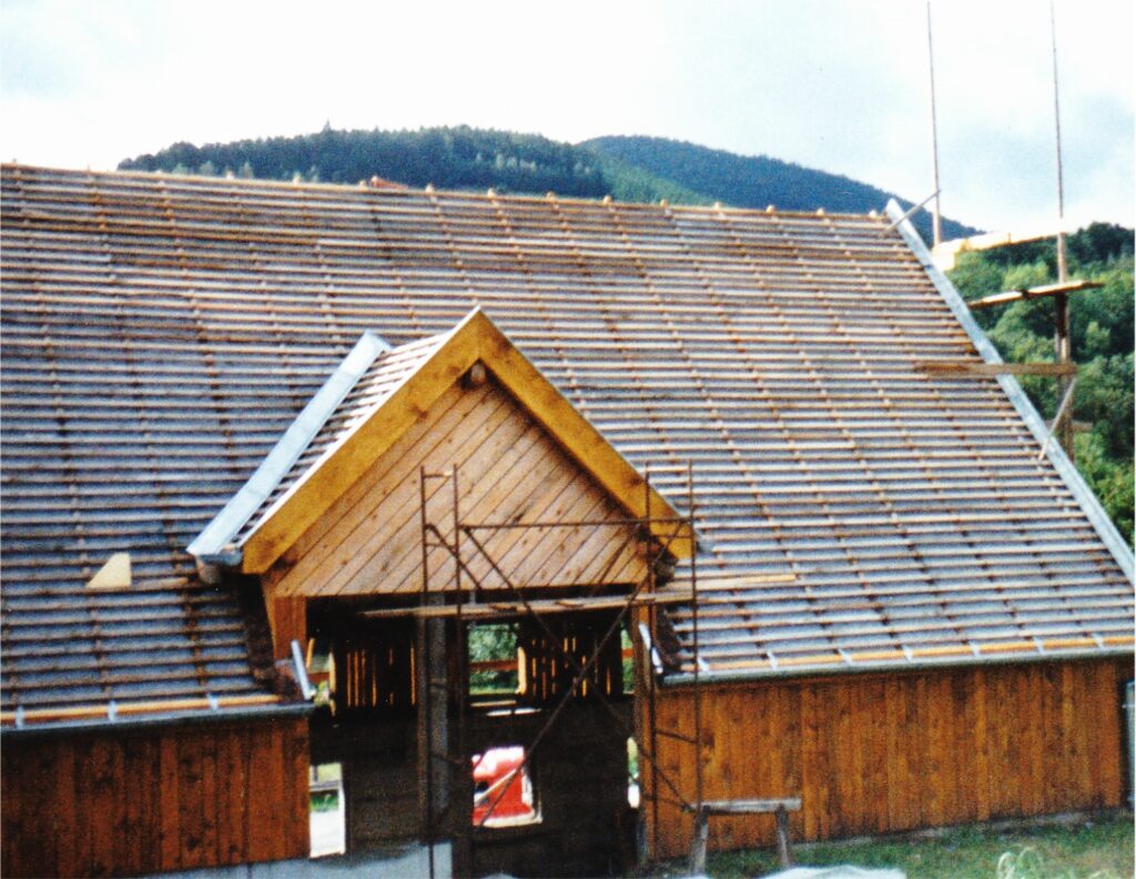 Wood-frame house under construction with a steep pitched wooden roof and scaffolding in a rural setting.