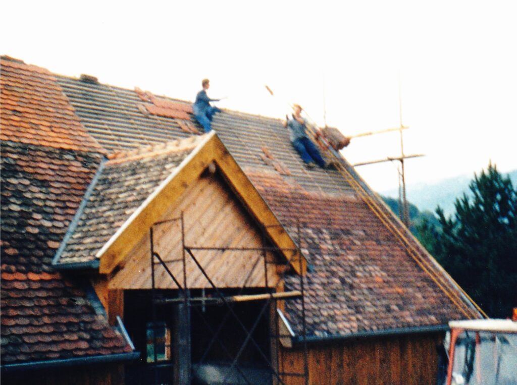 Two workers on a sloped roof laying tiles, scaffolding visible in front of a wooden house façade at a construction site.