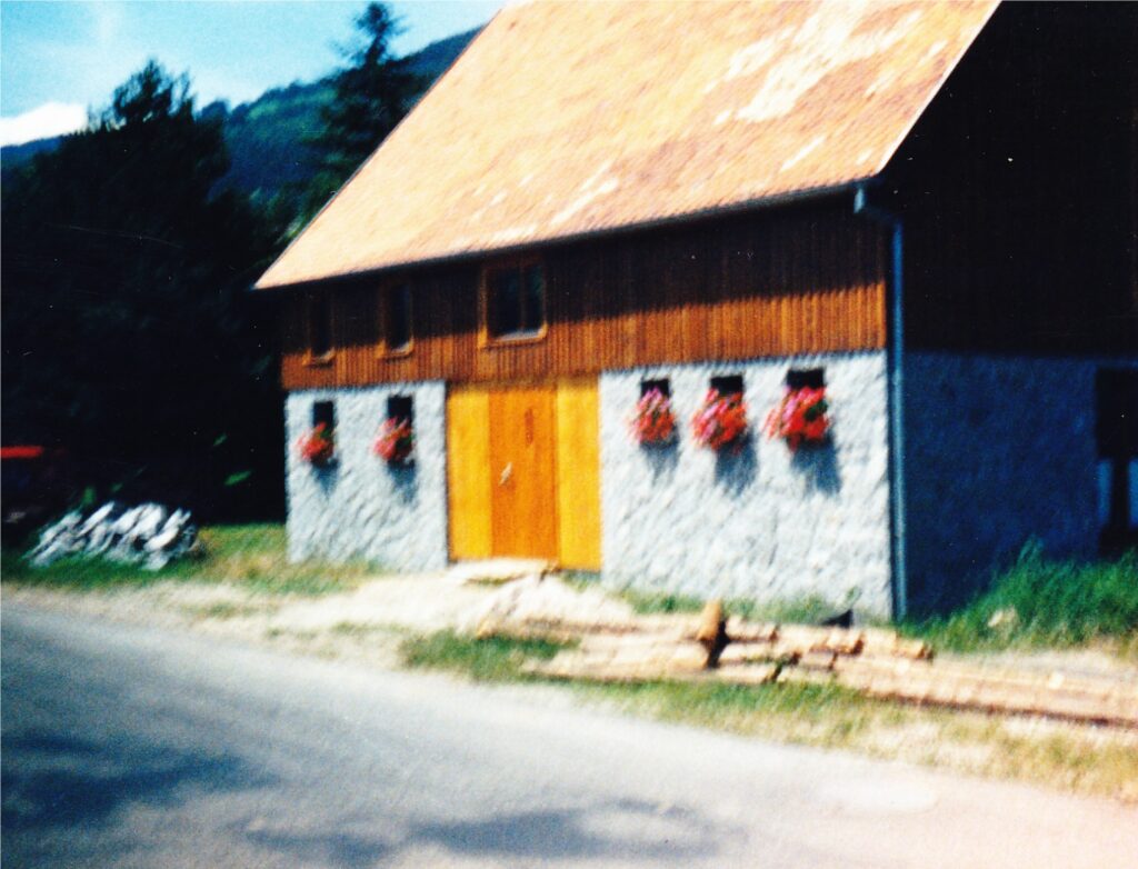 Rustic house with a stone base, wooden upper walls, orange doors, and window boxes of red flowers on a sunny day.