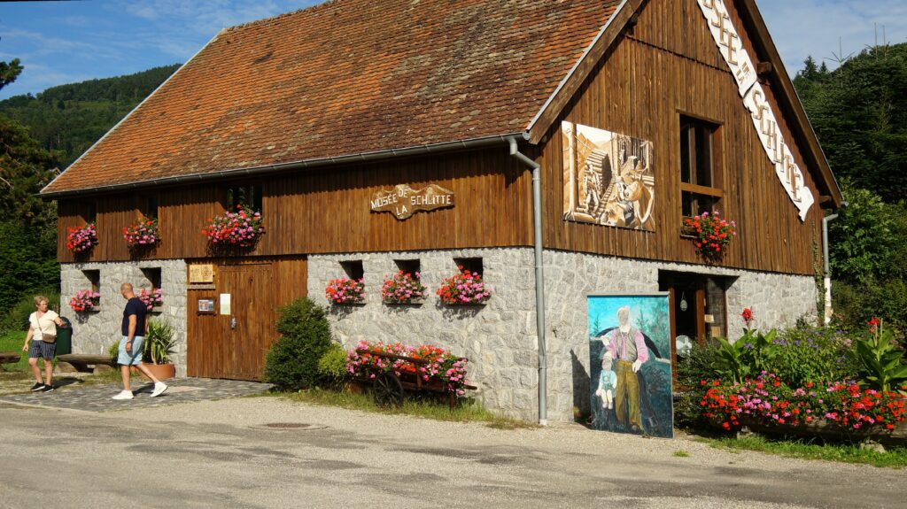 Wooden chalet-style museum with stone base, colorful flower boxes, and two visitors walking outside on a sunny day.