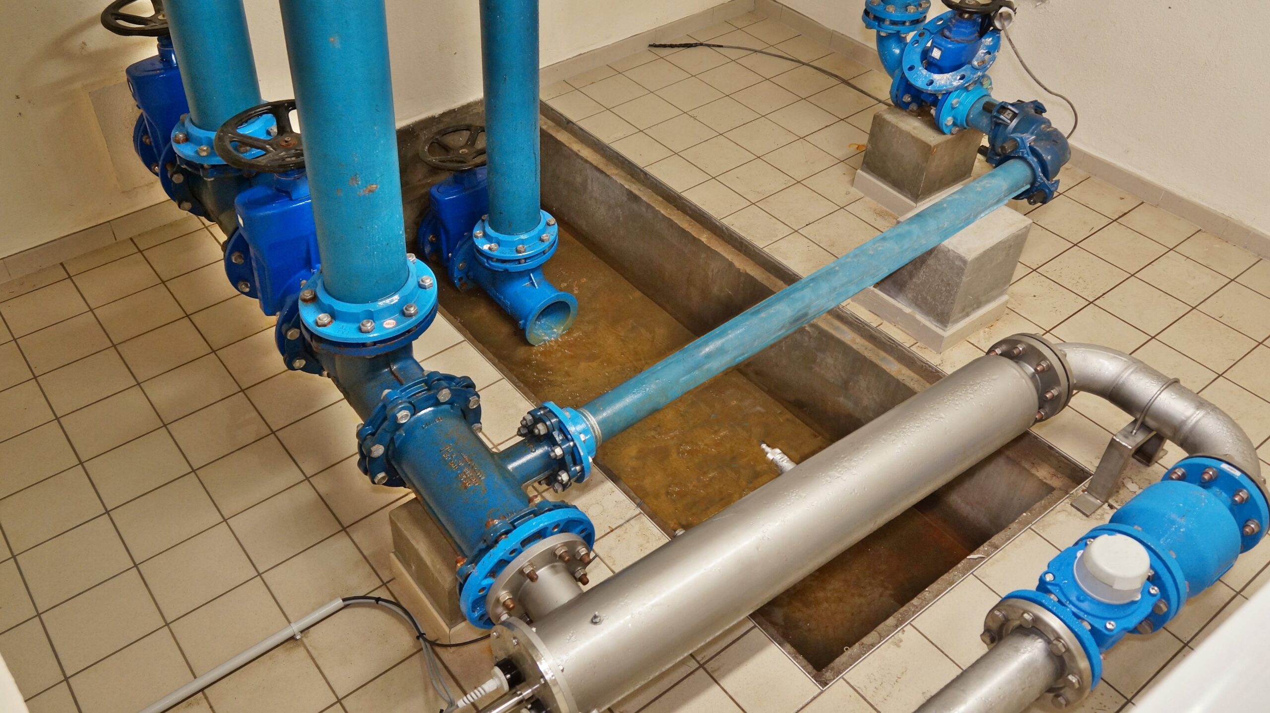 Blue piping system with flanged valves in a mechanical room or utility area, tiled floor visible.