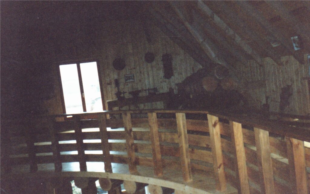 Dim attic with a curved wooden railing along a staircase, rough wooden walls, and light coming through double doors on the left.