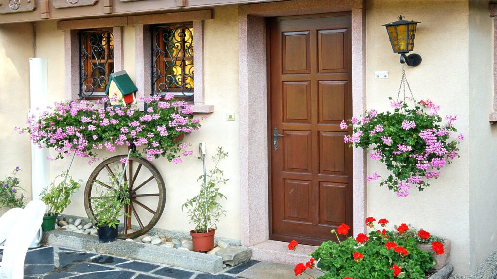 Front door entry of a beige house with a brown paneled door, lantern, and pink flowering pots beside a vintage wheel.
