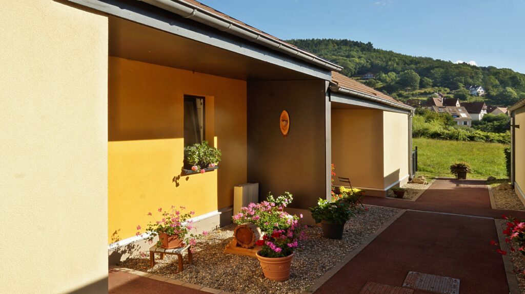 Sunny exterior of a yellow building with brown trim, along a gravel path lined with potted flowers.