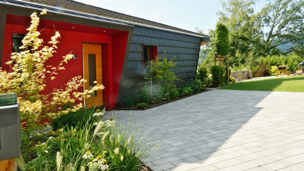 Modern house with a red entry and dark gray siding, garden plants in the foreground and a paved patio nearby.