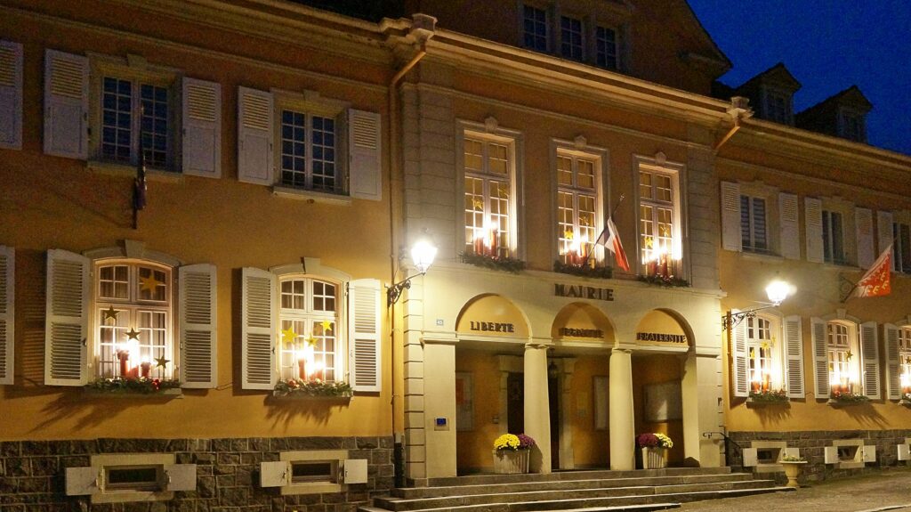 Night-time view of a French town hall (Mairie) with illuminated windows, flag, and star decor along a stone-paved street.”] ,