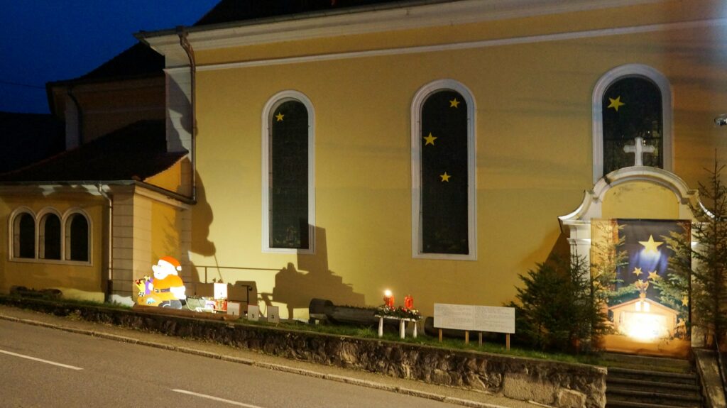 Nighttime view of a yellow church with arched windows and star decals in the windows, decorated for Christmas with a Santa figure and holiday banners outside