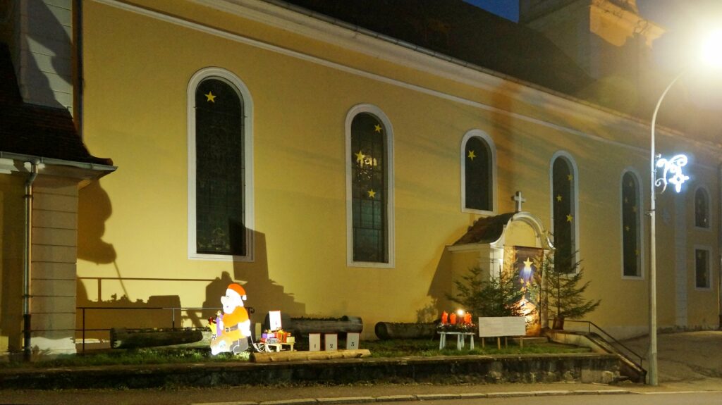 Night view of a yellow church with arched windows decorated with star stickers and Christmas decorations outside, including Santa figure and gifts.