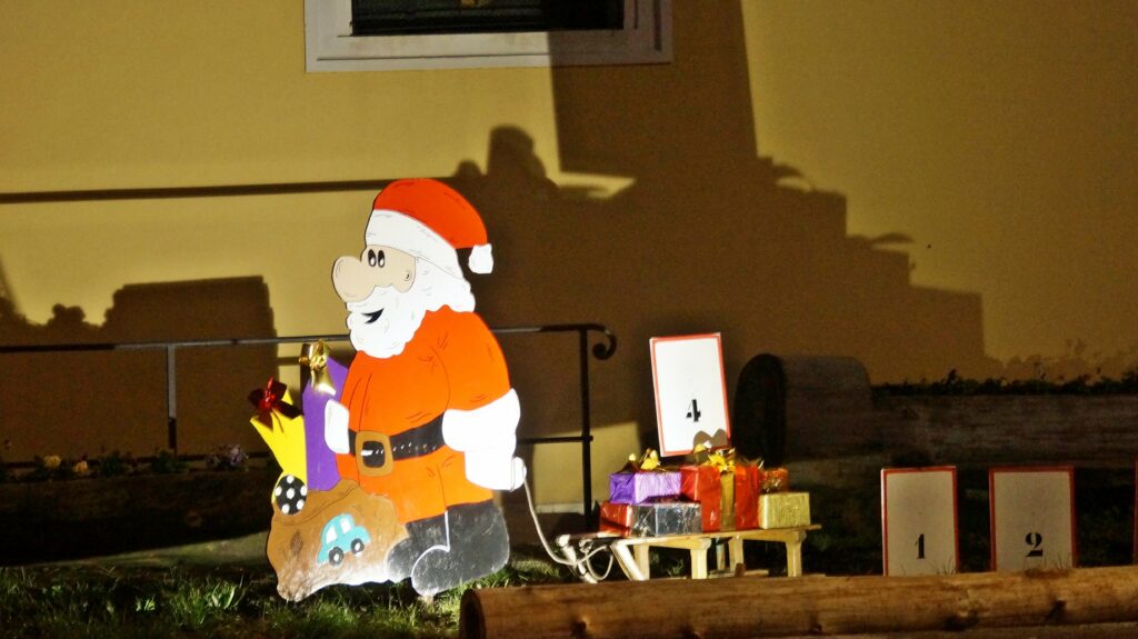 Life-size Santa cutout with a bag of gifts beside a stack of wrapped presents at an outdoor nighttime holiday display.
