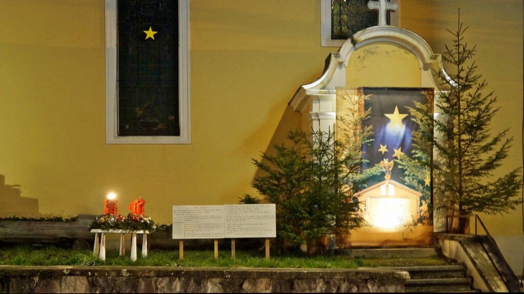 Night view of a yellow church wall with a window and a decorative arch, plus a starry banner and evergreen trees for a Christmas display outside the church.