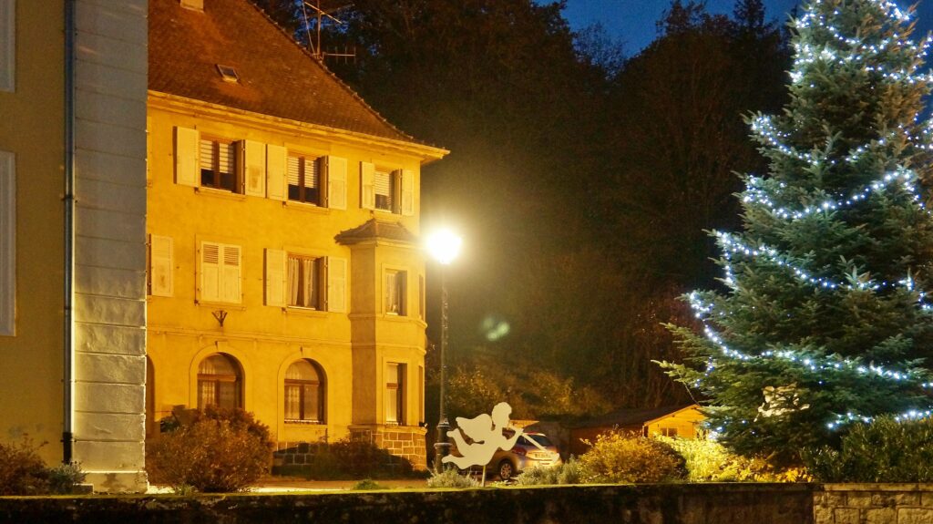 Night street scene featuring a yellow building with lit windows and a warmly lit street lamp, beside a tree with blue Christmas lights.