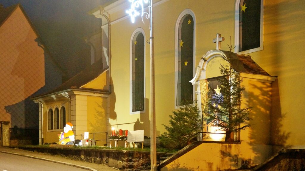 Night view of a yellow church facade with arched windows adorned by star decorations, and a Christmas display with a Santa figure, candles, and a small tree in front.