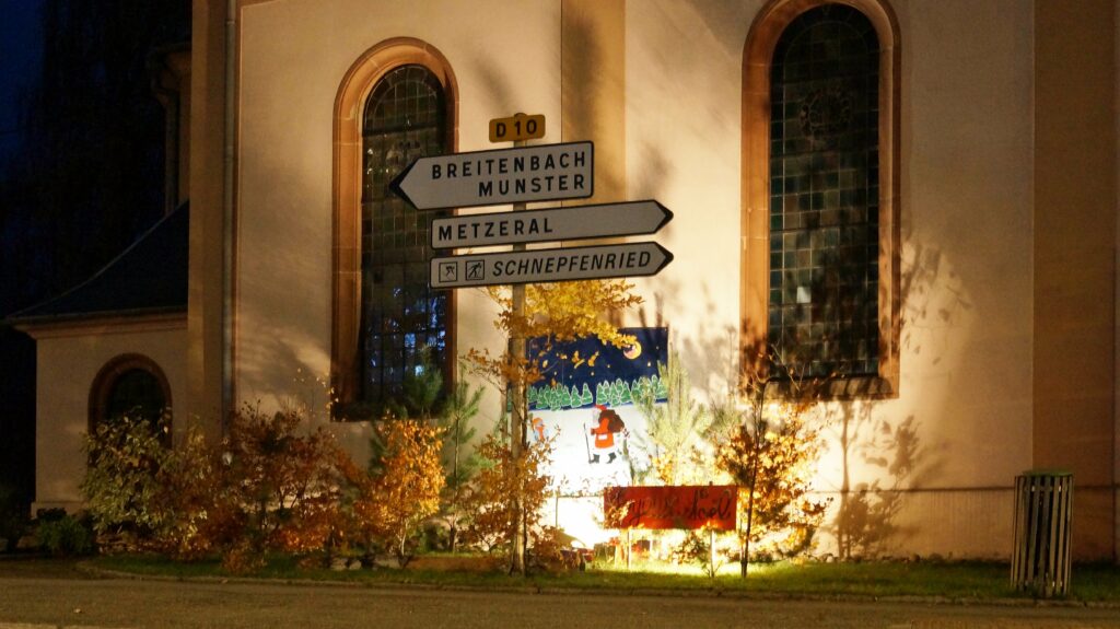 Night photo of a beige building with arched stained-glass windows and a four-direction sign reading Breitenbach, Münster; Metzéral; Schnepfénried.