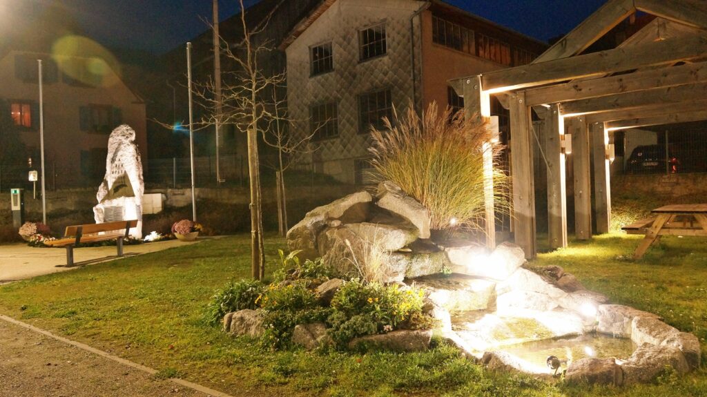 Night scene in a small park: a stone statue with a plaque, a wooden bench, rock garden, and warm lighting from a pergola structure.