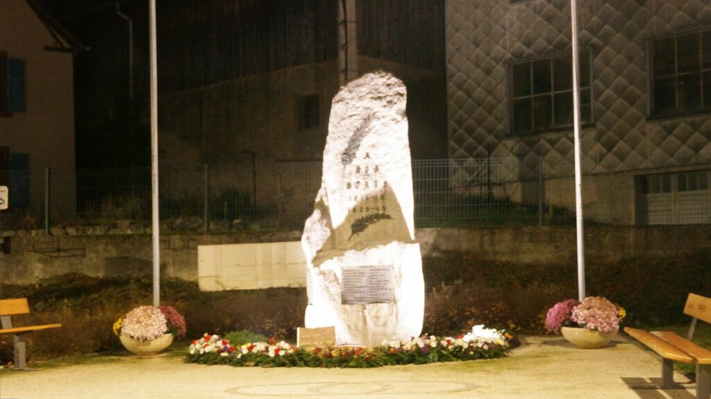 Night scene of a tall white stone monument surrounded by flower planters and benches in a plaza, with buildings in the background and spotlights on the monument.