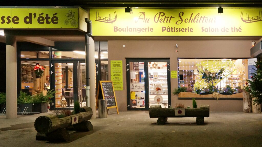 Night storefront of a French bakery and cafe with yellow signage reading 'Au Petit Schilteur' and 'Pâtisserie' visible, Christmas lights in window.