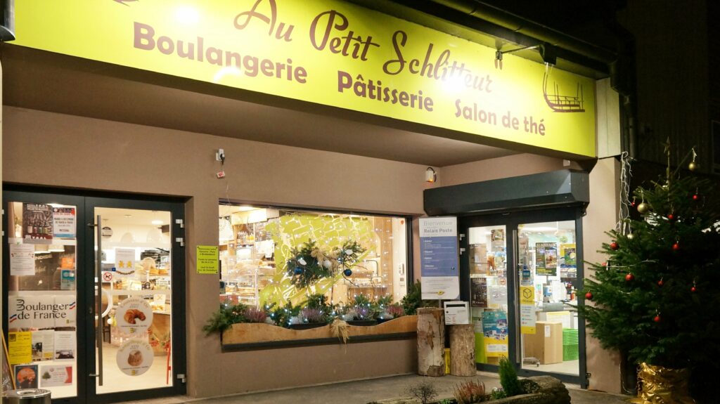 Front of a bakery storefront at night with a bright yellow sign and Christmas decorations outside. Visible glass doors and displays inside.