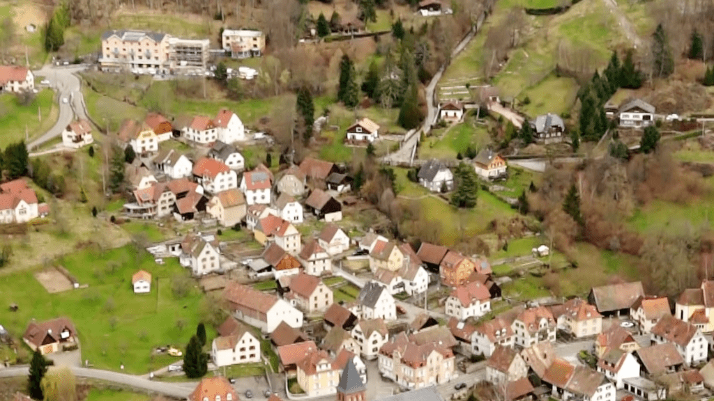 Aerial view of a small hillside village with many white houses and red roofs, winding roads and green fields interspersed among trees.