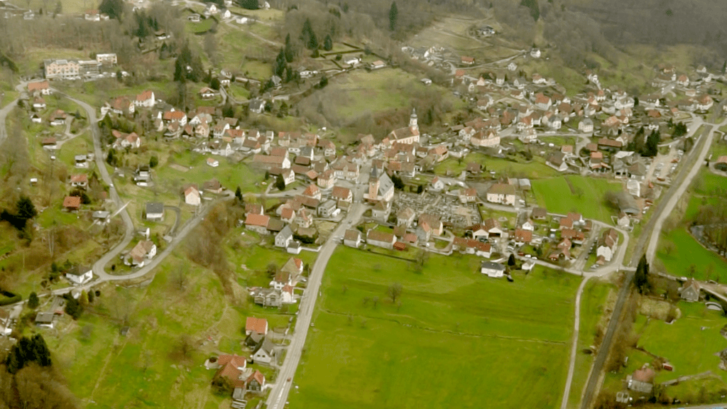 Aerial view of a small European village with a central church, red-roofed houses, and winding streets among green fields.