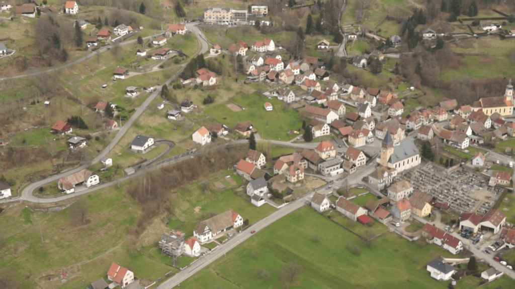 Aerial view of a small village with red-roofed houses, a prominent church with a tall tower, and winding roads across green fields.