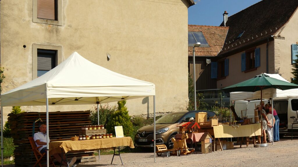 Outdoor market scene with white tents, wooden crafts and jars laid out on tables, people browsing and chatting.