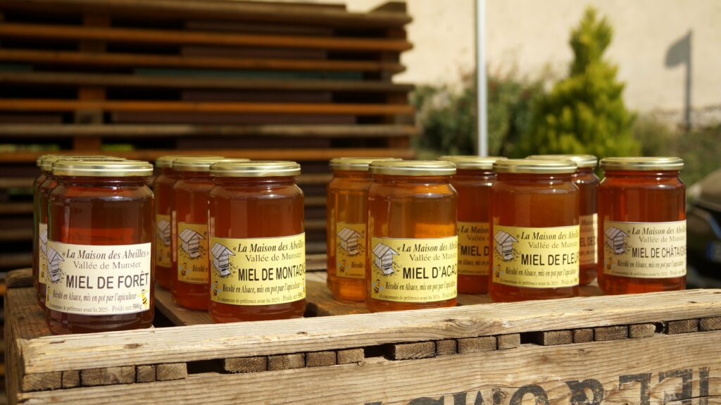 Row of honey jars with gold lids on a wooden pallet; labels read 'La Maison des Abeilles' and 'Miel de Forêt'