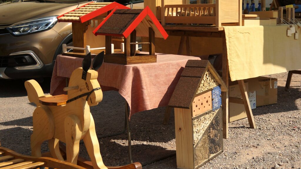 Outdoor market display of handmade wooden crafts: birdhouses, a wooden toy horse, and roof-panel pieces on a table covered with a pink cloth; a car is in the background.
