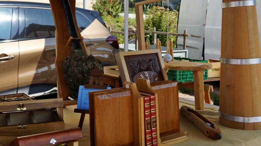 Outdoor market stall with wooden furniture, a carved clock, books, and barrels; a car is parked in the background.