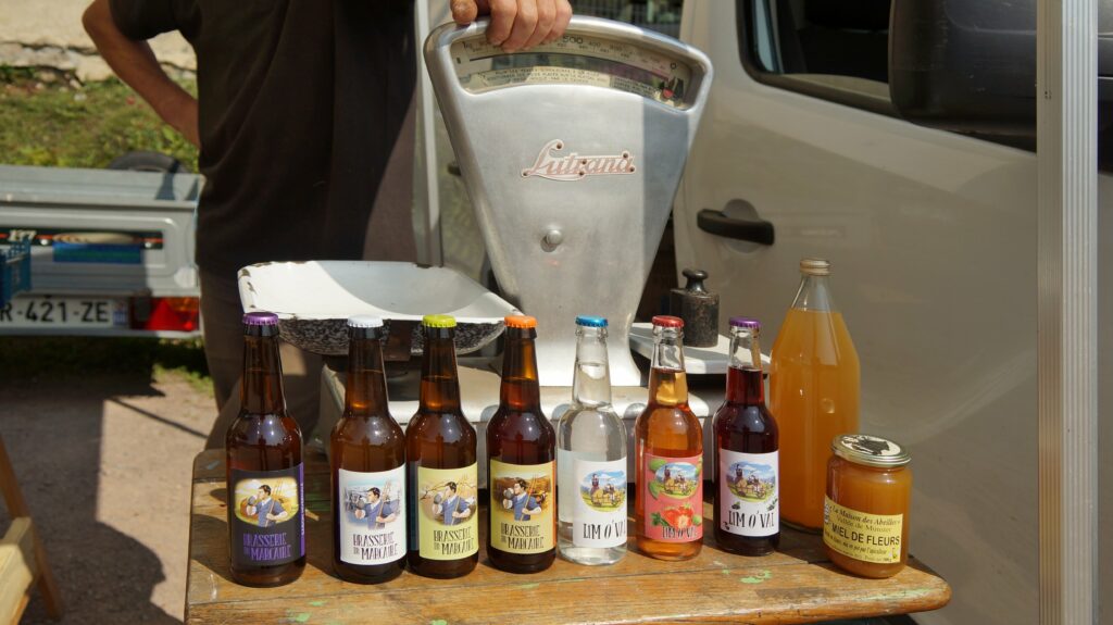 Row of glass bottles with colorful labels lined up on a wooden table beside a large metal vintage scale, outdoors at a market scene.