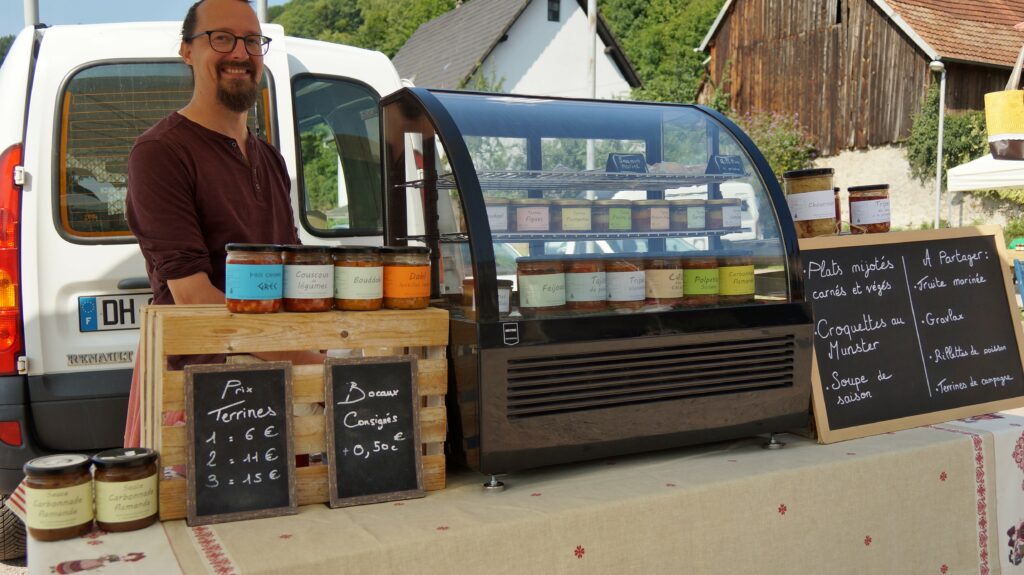 Vendor with glasses standing beside a glass display of labeled jars at an outdoor market. van in background and chalkboard menu nearby.