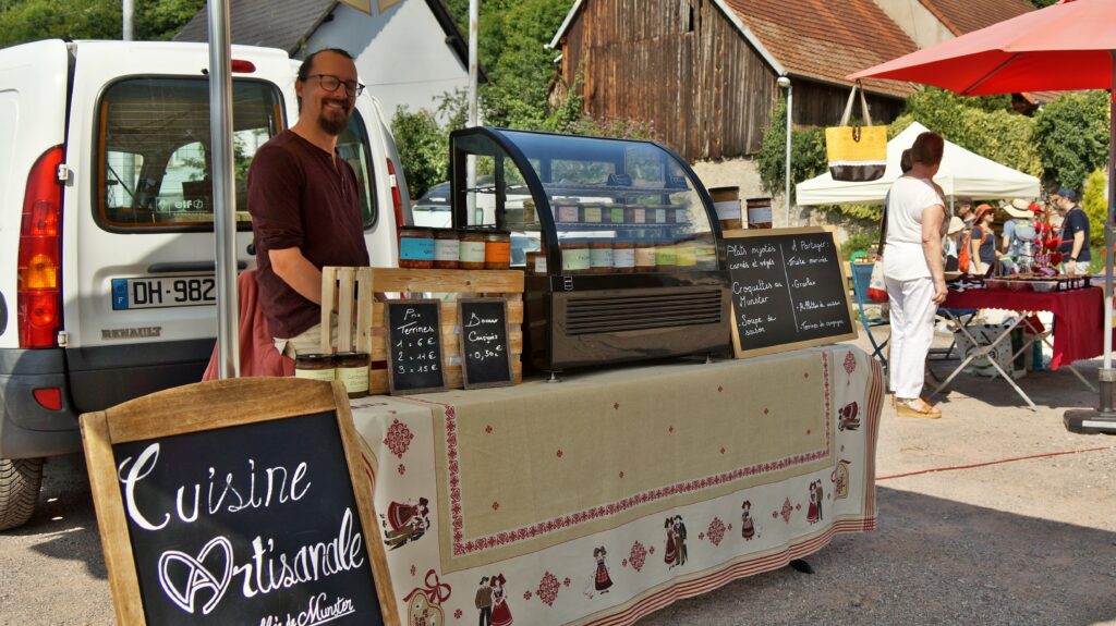 Vendor at an outdoor market stall with jars of jam and chalkboard prices, a white van parked behind the stand.