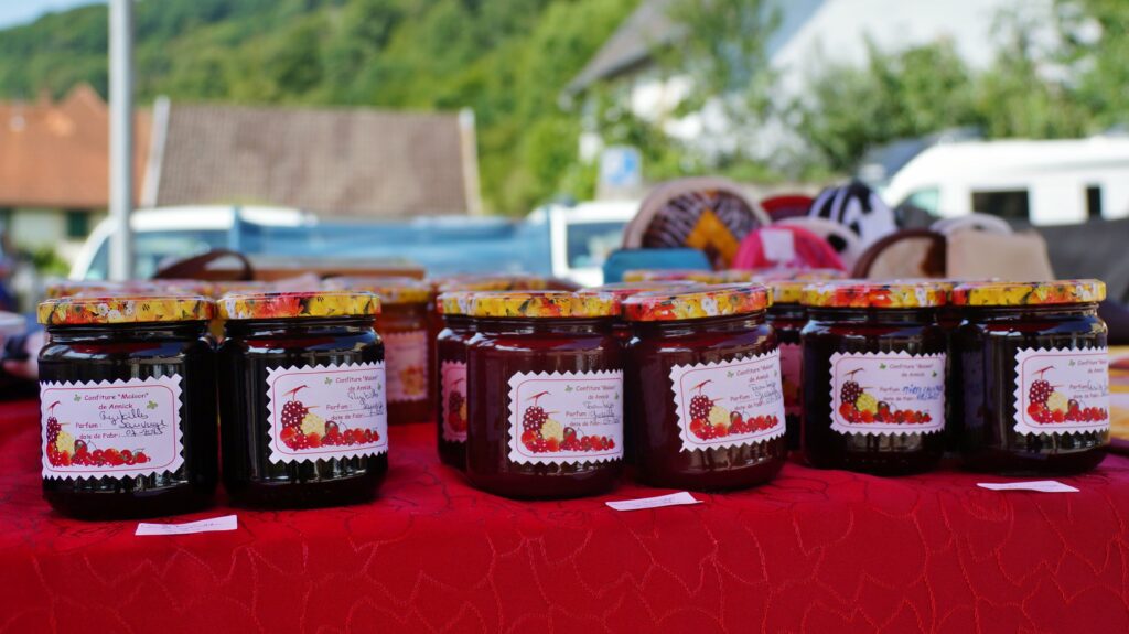 Row of glass jam jars with floral-labeled lids on a red table at an outdoor market stall