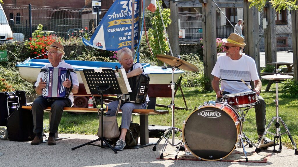 Three senior street musicians perform outdoors: two playing accordions and a drummer on the right, with a blue banner in the background.