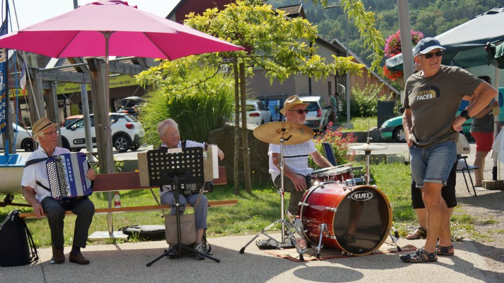 Outdoor street performance with three elderly musicians under a pink umbrella: accordion, drums, and a standing guitarist/singer.