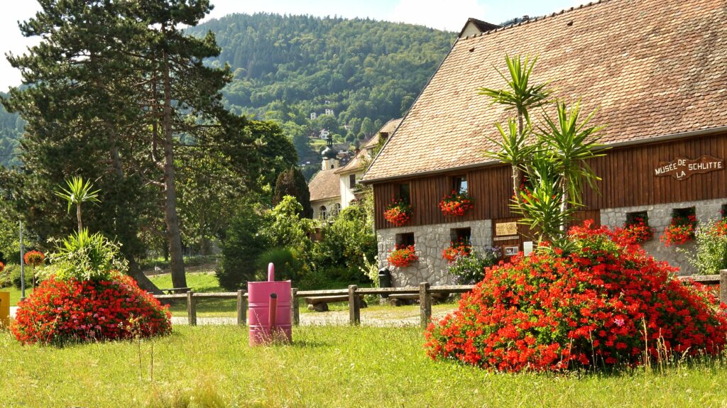 Rustic wooden museum building with red flower baskets, stone base, and lush garden in a rural valley.