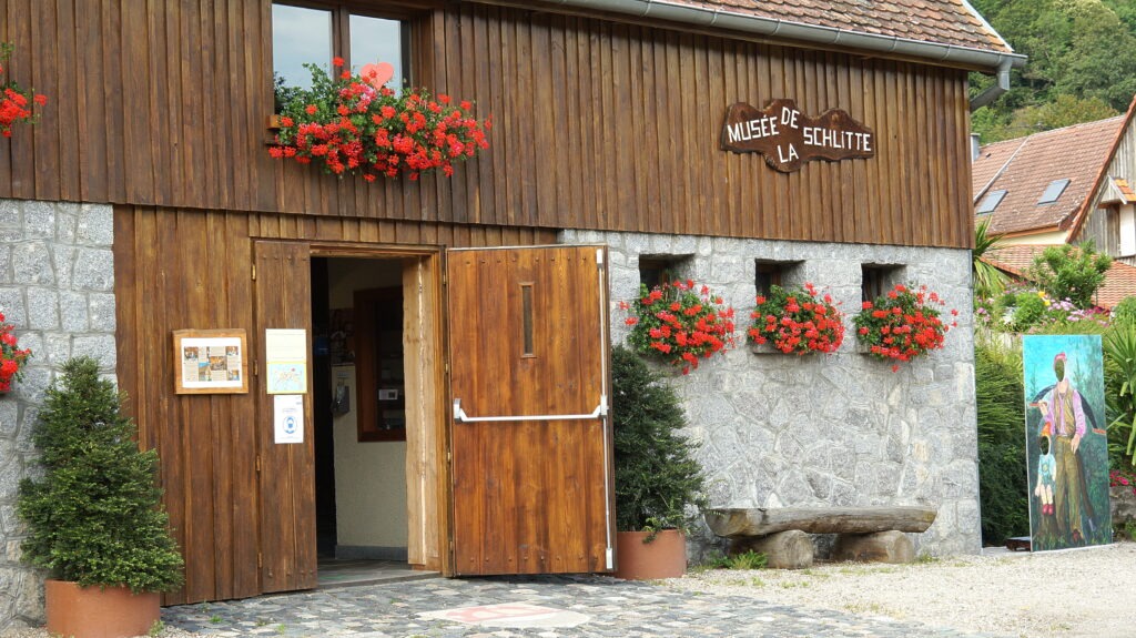 Wooden museum storefront with red flower boxes along a stone wall, open double doors inviting visitors.