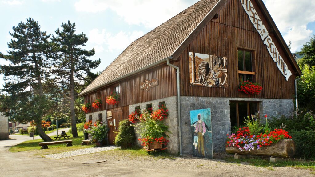 Rustic wooden museum building with a stone base, red flower boxes, and a mural on the wall amid a garden setting.