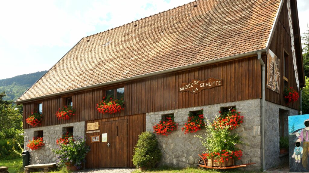 Wood-and-stone museum building with a steep wooden roof and red geranium window boxes, sign reads 'Musée de la Schlitte' on the facade.