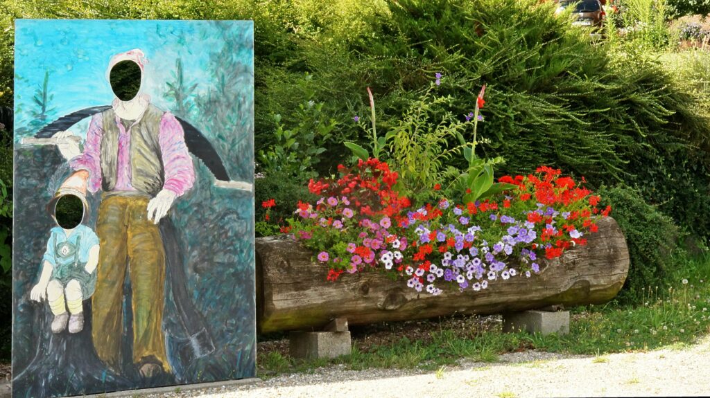 Outdoor mural: a man with a missing head and a child painted on a wooden panel, beside a flower-filled wooden planter.