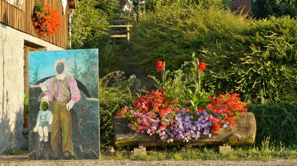 Outdoor garden scene with a painted panel depicting a man and child beside a colorful flower bed and wooden planter wall nearby.