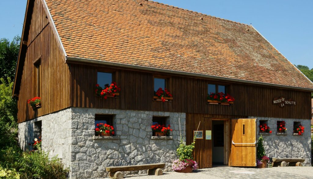 Rustic two-story building with a stone base, wooden upper walls, red flower boxes under windows, and open wooden doors; Musée de la Schlitte sign.