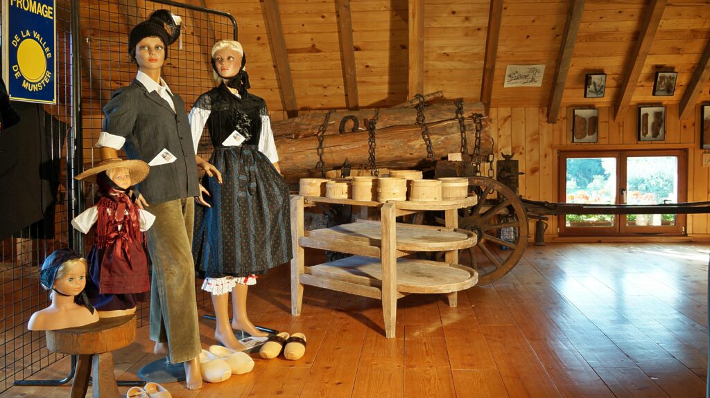 Life-size mannequins in vintage clothing (children and an adult) stand in a rustic museum setting with wooden walls, a wagon wheel, and stacked wooden buckets nearby.
